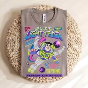 May include: A folded, grey t-shirt with a vibrant graphic of Buzz Lightyear from Toy Story. The design includes the words "Buzz Lightyear," "Space Ranger," and "To Infinity and Beyond." The shirt is displayed on a woven, circular mat.