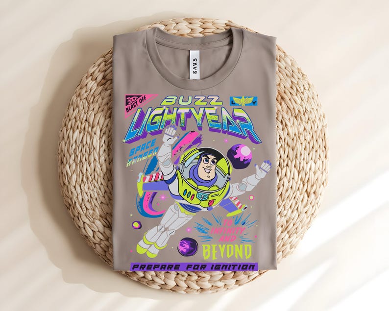 May include: A folded, grey t-shirt with a vibrant graphic of Buzz Lightyear from Toy Story. The design includes the words "Buzz Lightyear," "Space Ranger," and "To Infinity and Beyond." The shirt is displayed on a woven, circular mat.