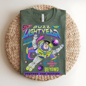 May include: Olive green t-shirt with a vibrant graphic of Buzz Lightyear in a spacesuit. The design includes the text "Buzz Lightyear", "To Infinity and Beyond", and "Prepare for Ignition". The shirt is displayed on a woven mat.