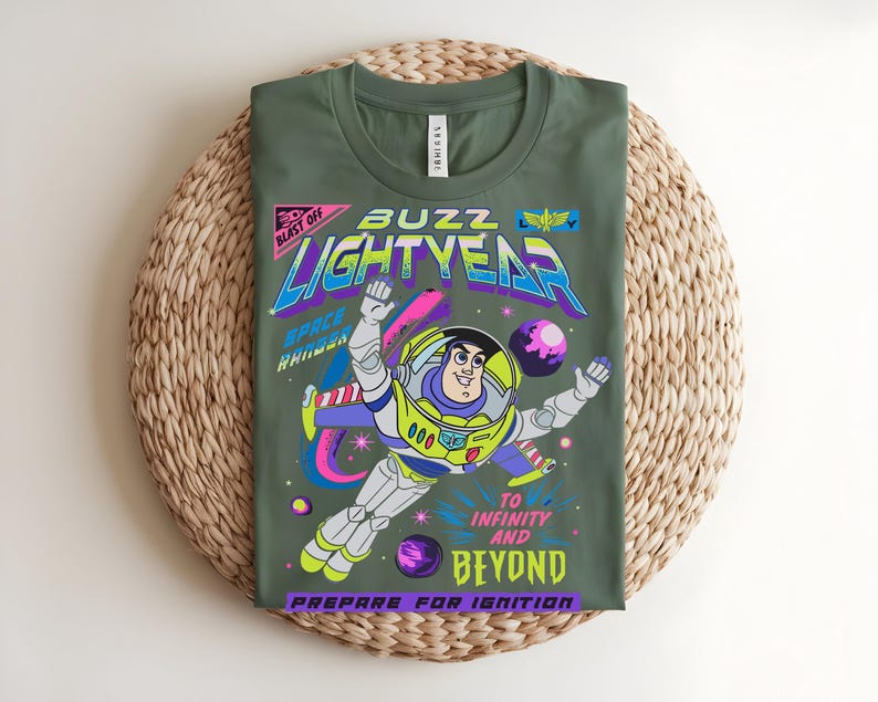 May include: Olive green t-shirt with a vibrant graphic of Buzz Lightyear in a spacesuit. The design includes the text "Buzz Lightyear", "To Infinity and Beyond", and "Prepare for Ignition". The shirt is displayed on a woven mat.