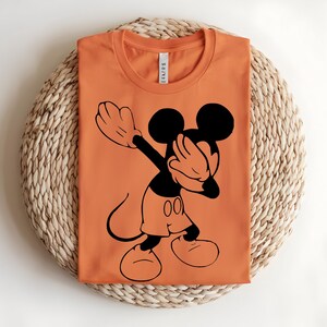 K&ouml;nnte beinhalten: Ein orangefarbenes T-Shirt mit Rundhalsausschnitt und einem schwarzen Grafikdruck einer Zeichentrickmaus, die eine Dab-Pose einnimmt. Das Shirt hat einen runden Halsausschnitt und ein wei&szlig;es Innenetikett, das am Kragen sichtbar ist.
