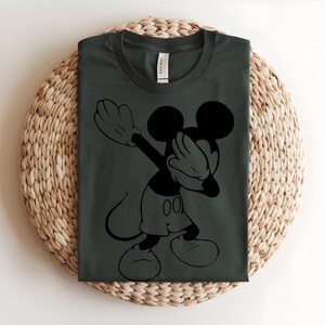 K&ouml;nnte beinhalten: Ein dunkel anthrazitfarbenes T-Shirt mit Rundhalsausschnitt und einem schwarzen Grafikdruck der Zeichentrickfigur Mickey Mouse, die eine Dab-Pose ausf&uuml;hrt. Das Shirt hat einen klassischen Kurzarm-Schnitt und ein sichtbares Markenetikett am Kragen.