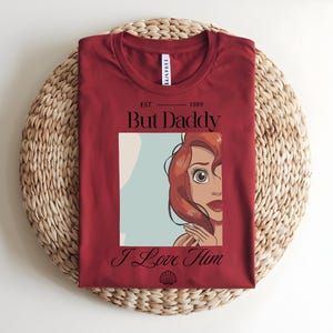 May include: A dark red t-shirt featuring a graphic print of a cartoon character with red hair. The text reads "EST 1989", "But Daddy", and "I Love Him" in black lettering. A small black seashell icon is printed below the text.