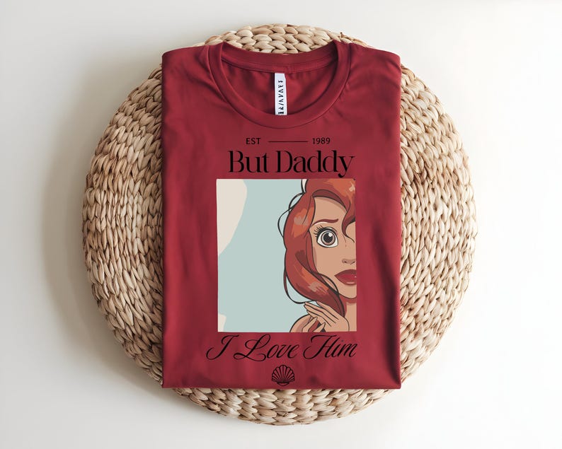 May include: A dark red t-shirt featuring a graphic print of a cartoon character with red hair. The text reads "EST 1989", "But Daddy", and "I Love Him" in black lettering. A small black seashell icon is printed below the text.