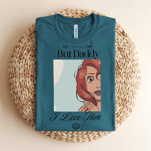 May include: A teal t-shirt featuring a graphic of a red-haired character with large eyes. The text reads "EST 1989", "But Daddy", and "I Love Him" in black lettering. A small black seashell icon is printed below the text on the front of the shirt.