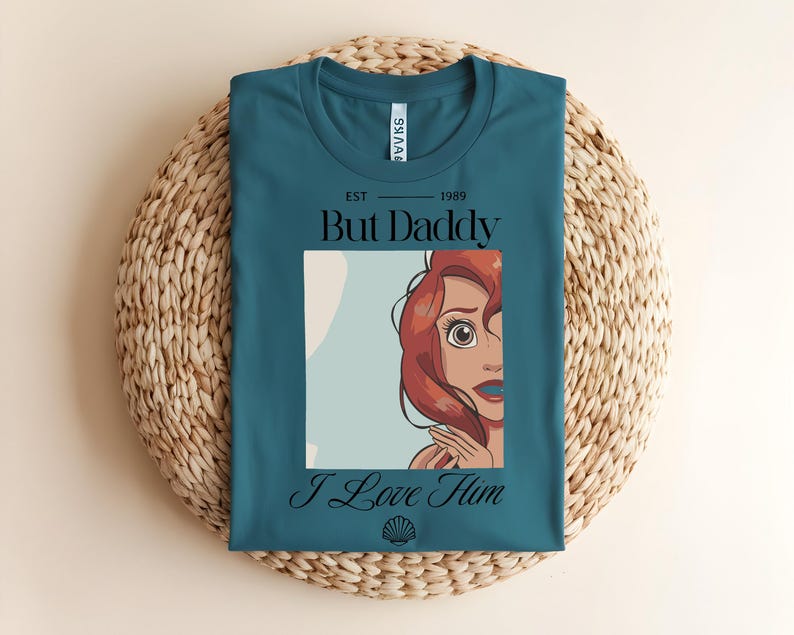 May include: A teal t-shirt featuring a graphic of a red-haired character with large eyes. The text reads "EST 1989", "But Daddy", and "I Love Him" in black lettering. A small black seashell icon is printed below the text on the front of the shirt.