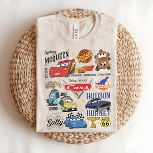 以下が含まれることがあります： ベージュ色のTシャツ。カラフルな漫画の車のグラフィックとテキスト要素が特徴です。デザインには、カーズの映画に登場するライトニング・マックィーン、メーター、ルイジ、グイド、サリーなどのキャラクターが含まれています。Tシャツは織られた表面に折りたたまれています。