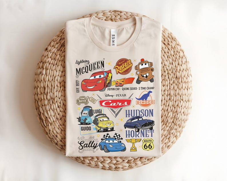 以下が含まれることがあります： ベージュ色のTシャツ。カラフルな漫画の車のグラフィックとテキスト要素が特徴です。デザインには、カーズの映画に登場するライトニング・マックィーン、メーター、ルイジ、グイド、サリーなどのキャラクターが含まれています。Tシャツは織られた表面に折りたたまれています。