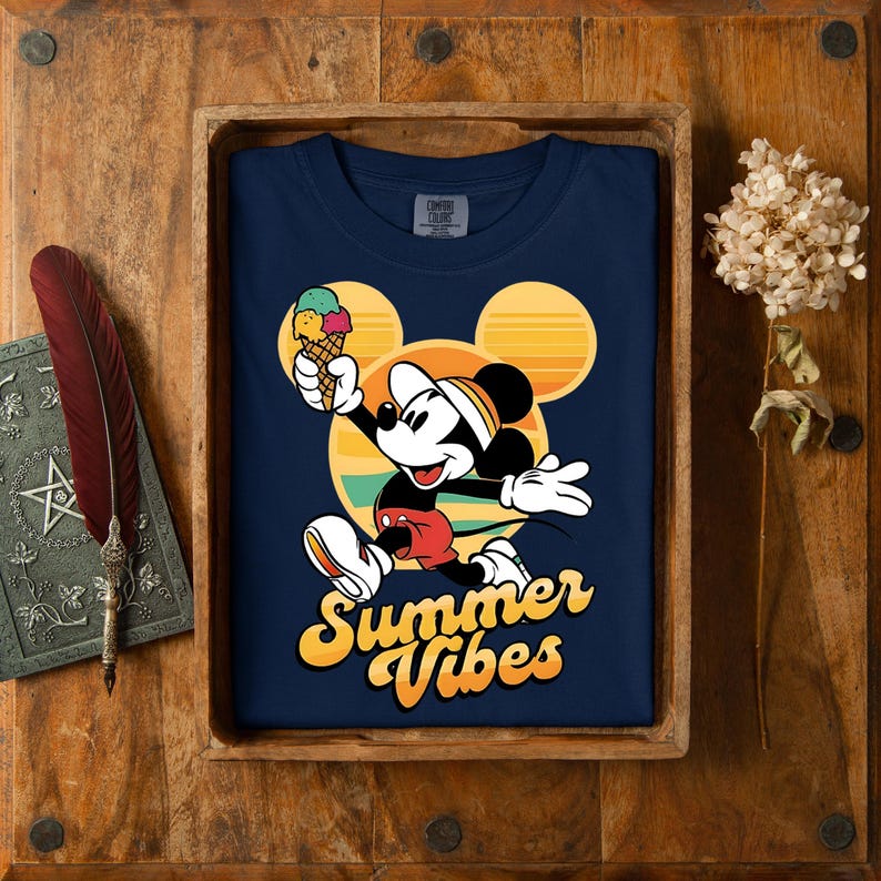以下が含まれることがあります： ミッキーマウスがアイスクリームを持っているネイビーブルーのTシャツ。デザインには、黄色とオレンジ色の「Summer Vibes」という文字が含まれています。Tシャツは木製のトレイに置かれ、羽と本が近くにあります。