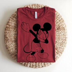 K&ouml;nnte beinhalten: Ein dunkelrotes T-Shirt mit Rundhalsausschnitt und einem schwarzen Grafikdruck von Mickey Mouse, der eine Dab-Pose ausf&uuml;hrt. Das Shirt hat ein wei&szlig;es Nackenetikett mit schwarzem Text am Kragen. Das Motiv ist mittig auf der Vorderseite platziert.