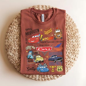 以下が含まれることがあります： さまざまな漫画の車のキャラクターと「Lightning McQueen」、「Cars」、および「Route 66」のテキストが特徴の、錆色のTシャツ。デザインには、レーシングカー、ロゴ、キャラクター名が含まれています。シャツは織りの表面に表示されます。