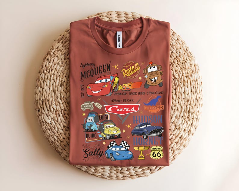 以下が含まれることがあります： さまざまな漫画の車のキャラクターと「Lightning McQueen」、「Cars」、および「Route 66」のテキストが特徴の、錆色のTシャツ。デザインには、レーシングカー、ロゴ、キャラクター名が含まれています。シャツは織りの表面に表示されます。