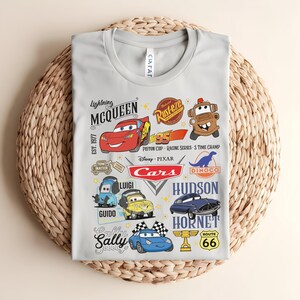 以下が含まれることがあります： 映画「カーズ」のキャラクターが描かれた、明るい灰色のTシャツ。ライトニング・マックィーン、メーター、その他のキャラクター、そして「Cars」と「Route 66」の文字がデザインされています。