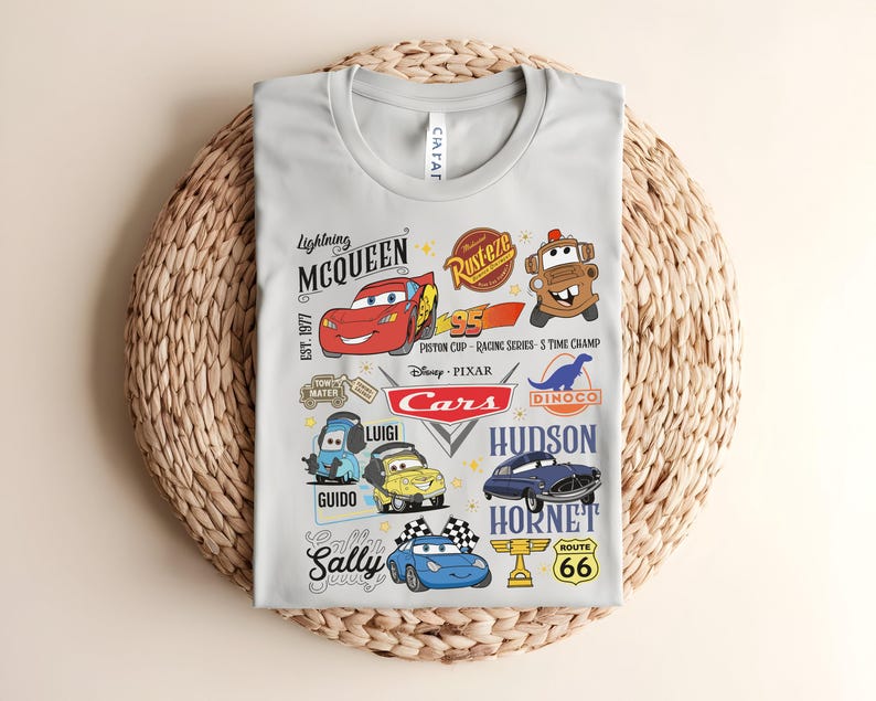 以下が含まれることがあります： 映画「カーズ」のキャラクターが描かれた、明るい灰色のTシャツ。ライトニング・マックィーン、メーター、その他のキャラクター、そして「Cars」と「Route 66」の文字がデザインされています。