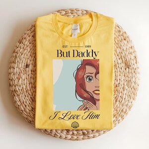 May include: A bright yellow t-shirt featuring a graphic of a red-haired character with large eyes. The text reads EST 1989 But Daddy I Love Him, with a small black seashell icon at the bottom. The shirt has a crew neckline and a white inner label.