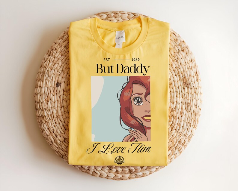 May include: A bright yellow t-shirt featuring a graphic of a red-haired character with large eyes. The text reads EST 1989 But Daddy I Love Him, with a small black seashell icon at the bottom. The shirt has a crew neckline and a white inner label.