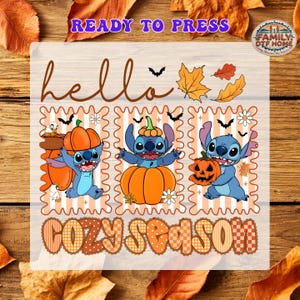 May include: A DTF transfer sheet featuring the text "hello" and "COZY SEASON" with three panels of a blue alien character interacting with orange pumpkins, autumn leaves, and small black bats on a light orange and white striped background.