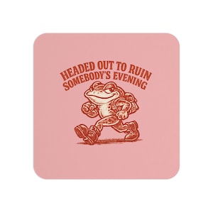 May include: A pink square coaster with a cartoon frog illustration in brown and white. The frog is walking and wearing a backpack. Text above the frog reads "HEADED OUT TO RUIN SOMEBODY'S EVENING".