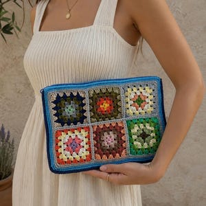 May include: A rectangular crochet clutch bag featuring a grid of six colorful granny squares. The squares display various yarn colors including blue, green, orange, yellow, pink, and brown, all bordered by a dark blue and light blue crocheted edge.