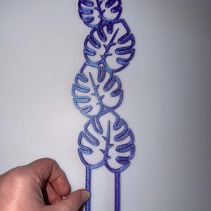 May include: A purple and blue glittery monstera leaf cake topper. The topper features five stylized leaves stacked vertically, with two prongs at the bottom for insertion into a cake. The leaves have a glittery finish.