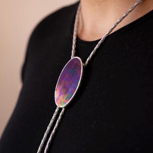 Puede incluir: Una corbata bolo con una pieza central ovalada de acabado iridiscente en tonos violeta y verde vibrantes. El colgante está enmarcado por un borde metálico y sujeto a un cordón trenzado de tono plateado que cuelga en dos hebras paralelas.