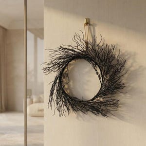 May include: A circular wreath constructed from thin, dark brown, twig-like branches. The wreath is suspended from a small, gold-coloured metal hook attached to a light-coloured wall. The structure features a dense, intricate arrangement of branching stems.