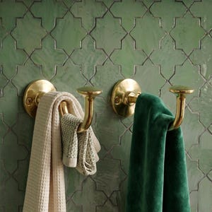 May include: Two polished brass wall hooks mounted on a green tiled wall. The left hook holds a cream-colored waffle-textured towel, while the right hook holds a solid dark green velvet towel. Each hook features a circular base and a curved arm.