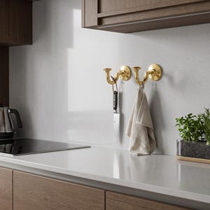 May include: Two polished brass wall hooks mounted on a light gray surface. The left hook holds a silver kitchen knife with a black handle by a leather loop, while the right hook holds a folded beige linen kitchen towel hanging vertically.
