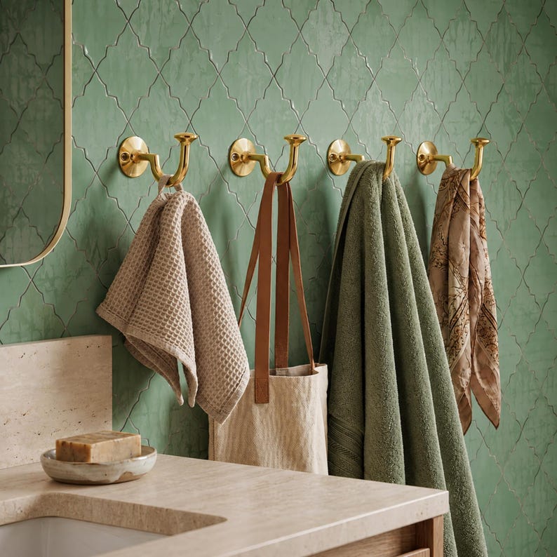 May include: Four polished brass wall hooks mounted in a row, each featuring a curved double-prong design. Hanging from the hooks are a beige waffle-weave towel, a tan tote bag with long straps, a sage green bath towel, and a patterned brown scarf.