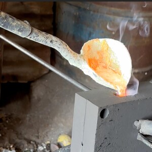 May include: A long metal ladle with a textured handle pours glowing orange molten metal into a rectangular gray mold. Wisps of white smoke rise from the point of contact where the liquid metal enters the mold, highlighting the casting process.
