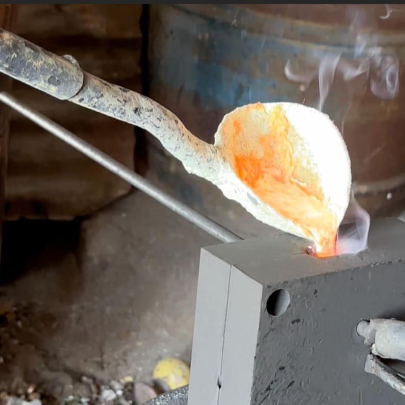 May include: A long metal ladle with a textured handle pours glowing orange molten metal into a rectangular gray mold. Wisps of white smoke rise from the point of contact where the liquid metal enters the mold, highlighting the casting process.
