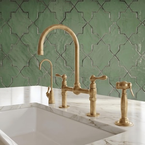 May include: A brushed brass kitchen faucet set featuring a high-arch spout, a separate side sprayer, and a smaller secondary faucet. The hardware is mounted on a white marble countertop with a green geometric tile backsplash behind the sink.
