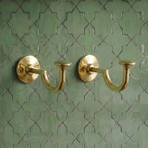 May include: Two polished brass curtain tieback hooks featuring a circular wall-mounting plate with two visible screws and a curved, J-shaped arm ending in a flat, circular finial. The hardware has a smooth, reflective metallic finish.