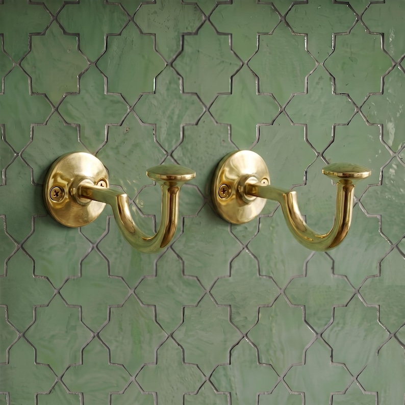 May include: Two polished brass curtain tieback hooks featuring a circular wall-mounting plate with two visible screws and a curved, J-shaped arm ending in a flat, circular finial. The hardware has a smooth, reflective metallic finish.