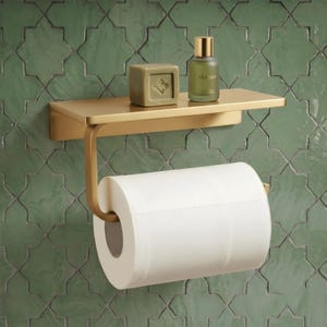 May include: A brushed gold metal toilet paper holder with an integrated flat shelf. A roll of white toilet paper hangs from the arm. A square beige soap bar and a small glass bottle with a gold cap labeled VEROANT sit on the top shelf.