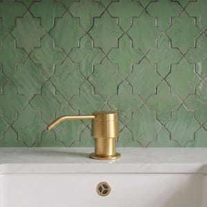 May include: A brushed gold soap dispenser with a cylindrical base and a slender, angled spout. The dispenser is positioned in front of a wall covered in sage green, star-shaped tiles with a glossy finish and dark grout lines.