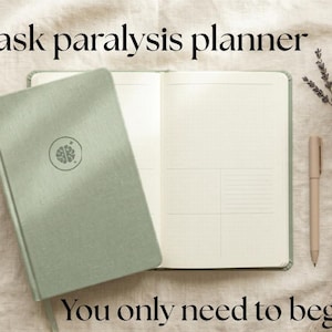 Könnte beinhalten: Ein offener, salbeigrüner Planer mit dem Text "ADHD Task paralysis planner" und "You only need to begin." Ein Stift, eine Tasse Tee und ein Pullover liegen ebenfalls auf einer cremefarbenen Oberfläche.