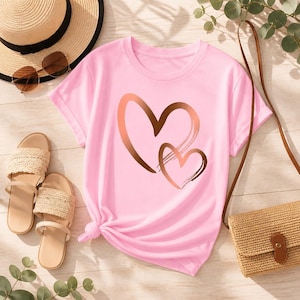 Könnte beinhalten: Hellrosa T-Shirt mit einem Design aus zwei sich überlappenden Herzen in Kupfer- und Brauntönen. Das Shirt ist an der Taille geknotet. Dazu ein Strohhut, Sonnenbrille, Sandalen und eine gewebte Handtasche.