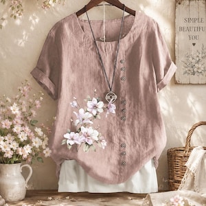 Könnte beinhalten: Staubrosa kurzärmlige Bluse mit floralem Aufdruck und Knopfleiste. Die Bluse hängt an einem Holzbügel. Eine lange Halskette mit rundem Anhänger ist sichtbar.
