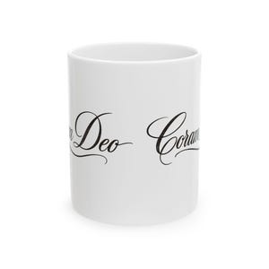 May include: A white ceramic mug featuring the black cursive text Coram Deo printed on the front. The mug has a cylindrical shape with a smooth finish and a simple handle-free design, showcasing the elegant script lettering clearly against the surface.