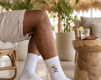 May include: White crew socks featuring a graphic of a yellow sun and dark blue palm trees with the text Rancho Colorados printed in a circular arc above the design. The socks are worn with white canvas sneakers and beige linen shorts.