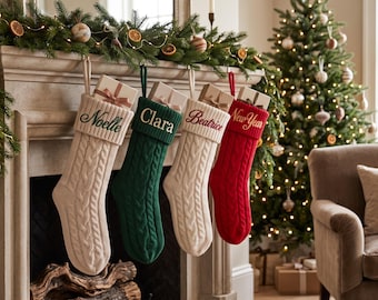 May include: Four cable-knit Christmas stockings hanging from a mantel, each featuring a ribbed cuff embroidered with a name: Noelle, Clara, Beatrice, and New Year. The stockings are cream, dark green, cream, and red, each holding a small gift box.