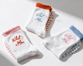 May include: Three pairs of white crew socks with ribbed cuffs in orange, pink, and light blue. Each pair features the embroidered text "What a Mom" in a matching color and a pattern of small heart-shaped grips on the sole and heel area.