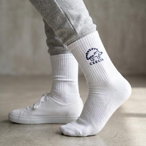 May include: A pair of white crew socks featuring a dark blue embroidered graphic of a hedgehog and the text CEECIL on the outer side of the ankle. The socks have a ribbed texture and are worn with grey sweatpants and white sneakers.