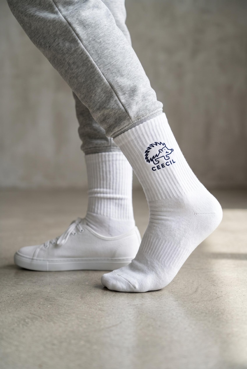 May include: A pair of white crew socks featuring a dark blue embroidered graphic of a hedgehog and the text CEECIL on the outer side of the ankle. The socks have a ribbed texture and are worn with grey sweatpants and white sneakers.