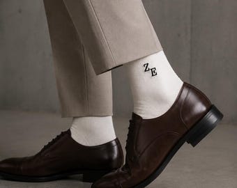 May include: A pair of brown leather dress shoes with black soles and laces. The shoes are paired with white socks featuring the embroidered letters "ZE" and beige trousers. The image is shot from a low angle.