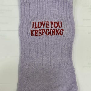 May include: A light lavender ribbed crew sock featuring the embroidered text "I LOVE YOU KEEP GOING" in a dark red thread. The sock has a vertical ribbed texture extending from the cuff down to the heel area, with a clear, legible font.