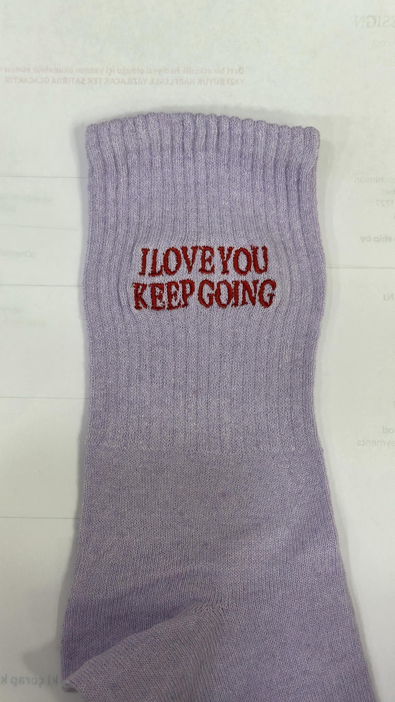 May include: A light lavender ribbed crew sock featuring the embroidered text "I LOVE YOU KEEP GOING" in a dark red thread. The sock has a vertical ribbed texture extending from the cuff down to the heel area, with a clear, legible font.