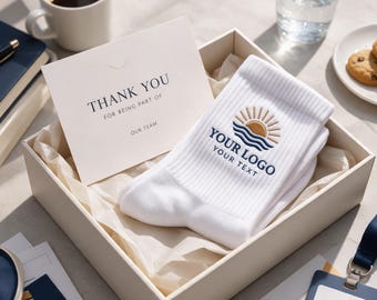 May include: A white pair of ribbed socks featuring an embroidered logo of a golden sun rising over blue waves, with the text "YOUR LOGO" and "YOUR TEXT" below it, presented in a gift box alongside a card that reads "THANK YOU FOR BEING PART OF OUR TEAM."