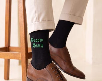 May include: Black crew socks featuring the text Groom Gang embroidered in green gothic lettering. The socks are worn with brown leather oxford shoes and light beige trousers. The footwear is styled with a classic cap toe and thin brown laces.